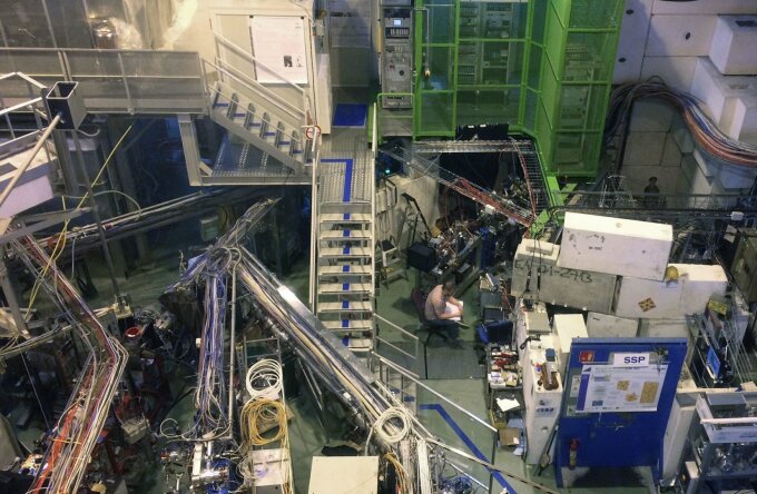 Versuchsaufbau in der ISOLDE-Halle. Hinter dem Forscher befindet sich der Versuchsaufbau, in dem die Astat-Ionen mit den Laserstrahlen überlagert werden. Die Laser befinden sich in dem grauen Raum auf der ersten Etage des Treppenaufgangs. Der Forscher überwacht gerade mit einem Oszil¬loskop die vom Detektor kommenden Signale der auftreffenden neutralen Astat-Atome und macht dazu Notizen in das analoge Logbuch.