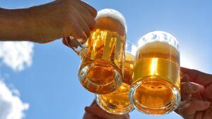 Three hands can be seen in front of a blue sky, each toasting with a glass of beer.