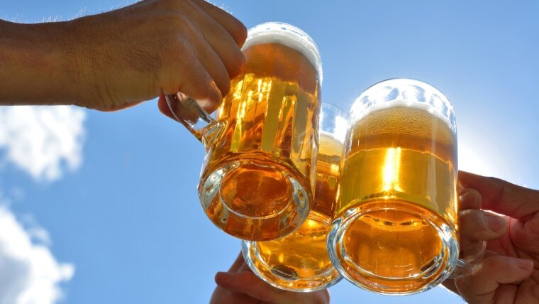Vor einem blauen Himmel sind drei Hände zu sehen, die mit jeweils einem Glas Bier anstoßen.